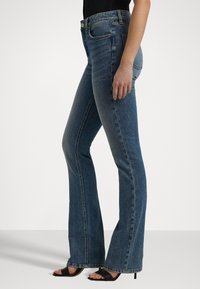 Jeans de mezclilla azul acampanados con una cintura de media altura, ligera desgastación y cinco bolsillos. Textura suave con un corte clásico y costuras cosidas.