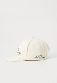 Cream-colored cap with a curved brim, featuring green embroidered text on the front and sides, made of cotton fabric.