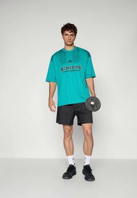 Vyras, vilkintis mėlyną Adidas sportinę marškinių ir juodas šortus, laiko svarmenų plokštę prieš paprastą baltą fono.