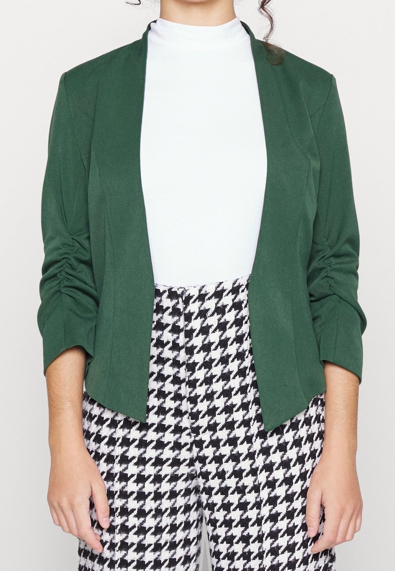 Zelený blejzr s mírně strukturovaným designem, dlouhými rukávy a nabíranými detaily. Spárovaný s kalhotami s černobílým kostkovaným vzorem.