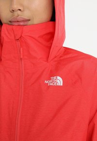 Veste à capuche rouge avec une finition texturée, dotée d'une fermeture éclair sur le devant et du logo North Face sur le côté gauche de la poitrine.