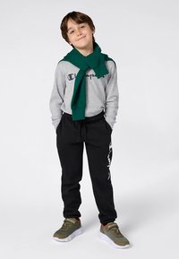 Graues Langarmshirt mit "Champion"-Logo, dunkelgrüner Pullover um den Hals gebunden, schwarze Jogginghose und olivfarbene Sneakers mit weißen Sohlen.