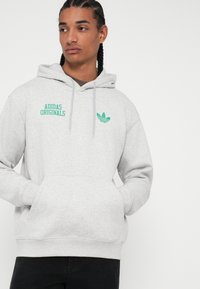 Szara bluza z kapturem wykonana z miękkiego materiału, z zielonym napisem i logo "Adidas Originals" na piersi, z kieszenią typu kangaroo i kapturem ze sznurkiem.
