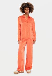 Donna con capelli ricci che indossa una camicia arancione lucida e larga con bottoni e pantaloni coordinati a gamba larga, in piedi con un'espressione neutra e sneakers.