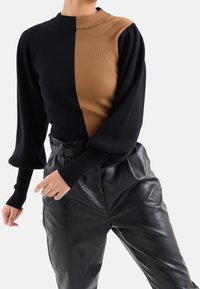 Osoba w prążkowanym swetrze w czarne i brązowe pionowe połowy oraz w czarnych skórzanych spodniach z wysokim stanem na białym tle.