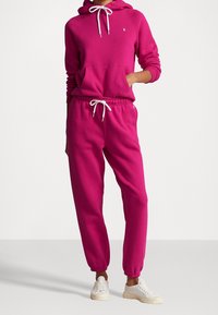 Rosa luvtröja med framficka, matchad med rosa mjukisbyxor. Båda har elastiska muddar och dragsnören. Vita sneakers ingår.