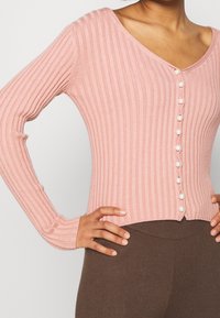 Różowy prążkowany kardigan z dekoltem w serek, z rzędami guzików przypominających perły z przodu i długimi rękawami. Miękka faktura i dopasowany krój.