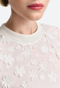 Weißes Oberteil mit floralem Muster und strukturiertem Design, versehen mit dreidimensionalen Blumen und Strass-Akzenten. Rundhals-Ausschnitt in cremiger Ribben-Optik.