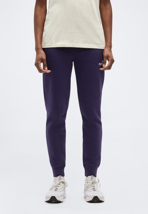 Personne portant un pantalon de survêtement Puma bleu marine, un t-shirt beige et des baskets blanches Puma, debout devant un fond uni.