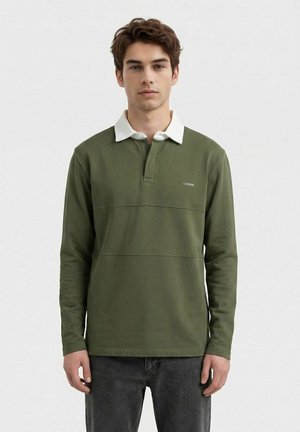 Ung mann med kort brunt hår iført en olivengrønn langermet polotrøye med hvit krage og mørke jeans, stående foran en ensfarget bakgrunn.