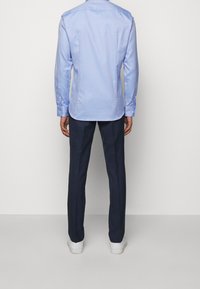 Camicia azzurro chiaro a maniche lunghe con polsini a bottone, yoke posteriore e orlo curvo; abbinata a pantaloni blu scuro e sneakers bianche.