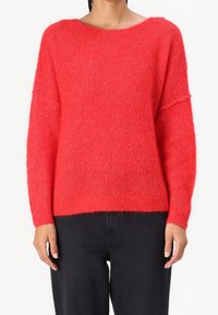 Pull rouge en maille avec une coupe ample, un large décolleté et des manches longues. Texture douce et ourlet côtelé. Porté avec un pantalon noir.
