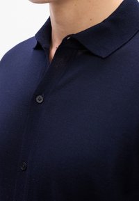 Gros plan d'une personne portant un polo bleu marine boutonné avec une texture tricotée douce et un col plié.