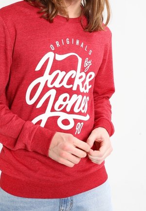 Person wearing a red sweatshirt with white "Jack & Jones Originals 90" text, adjusting sleeves, against a plain white background.