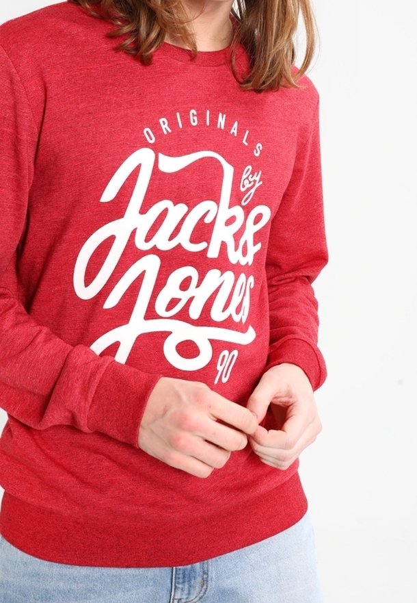 Persona che indossa una felpa rossa con la scritta bianca "Jack & Jones Originals 90", mentre sistema le maniche, su uno sfondo bianco uniforme.