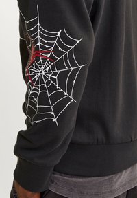 Sudadera negra con un diseño de telaraña blanca y una araña roja en la manga izquierda; puños y dobladillo acanalados para mayor textura.