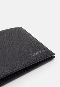 Portofel negru din piele, cu suprafață texturată, având un logo Calvin Klein vizibil, cu un finisaj metalic subtil. Design compact cu pliere.