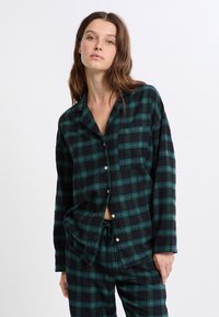 Kobieta ubrana w zieloną i czarną koszulę nocną w kratę oraz pasujące spodnie, stojąca na tle jasnym.