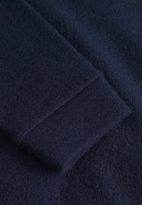 Temno modra volnena tkanina z mehkimi teksturami, ki prikazuje skrbno obdelan detajl manšete. Material izgleda debel in topel.