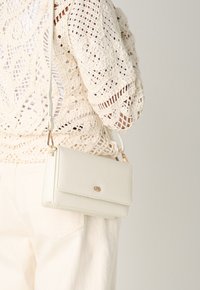 Borsa a tracolla bianca con una texture liscia, hardware oro e chiusura a pattina, indossata su uno sfondo di un indumento all'uncinetto beige.