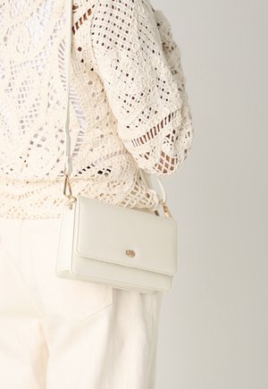 Weißes Umhängetasche mit glatter Oberfläche, goldenen Beschlägen und einem Klappverschluss, getragen vor einem Hintergrund eines beigen gehäkelten Kleidungsstücks.