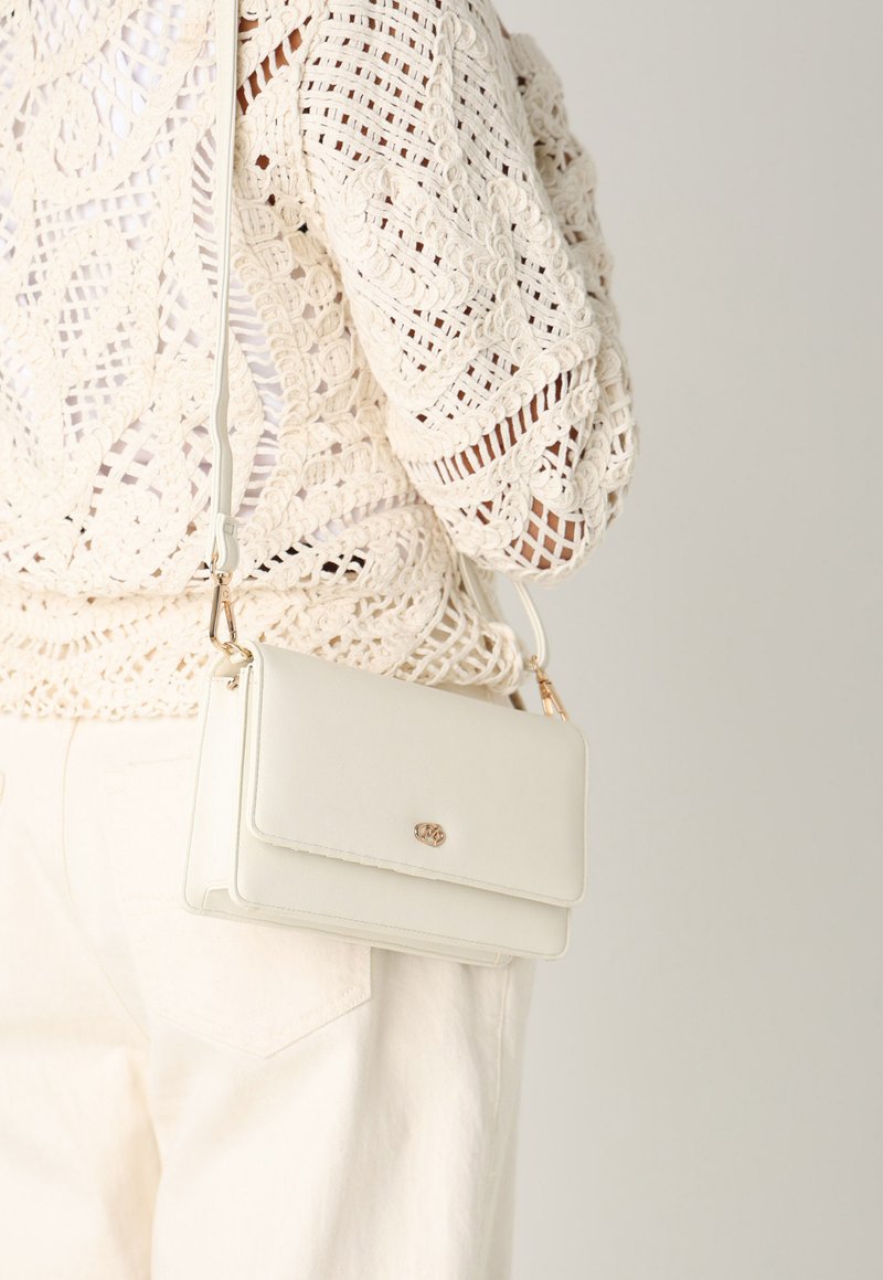 Borsa a tracolla bianca con una texture liscia, hardware oro e chiusura a pattina, indossata su uno sfondo di un indumento all'uncinetto beige.