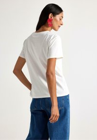 Biała bawełniana koszulka z krótkim rękawem, luźny krój, połączona z ciemnoniebieskimi luźnymi dżinsami. Modelka nosi dużą biżuterię w czerwone kwiaty.