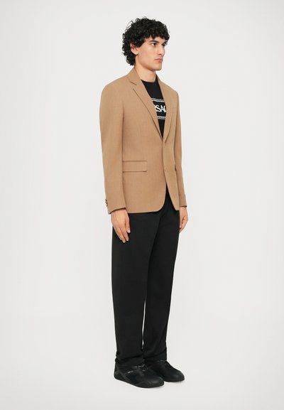 Blazer marrón hecho de tela texturizada, con dos bolsillos frontales, usado sobre una camiseta negra y combinado con pantalones negros y zapatillas.