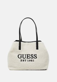Sac fourre-tout en fausse fourrure crème avec des poignées en cuir noir. Présente "GUESS" et "EST 1981" brodés en noir à l'avant.