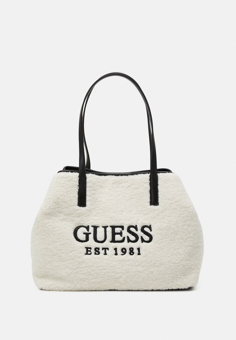 Sac fourre-tout en fausse fourrure crème avec des poignées en cuir noir. Présente "GUESS" et "EST 1981" brodés en noir à l'avant.