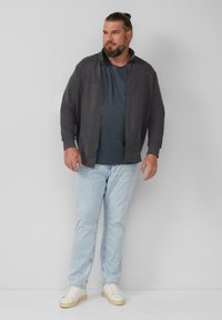 Dunkelgraue Jacke mit Reißverschluss über einem marineblauen T-Shirt, hellblauen Jeans und weißen Sneakers. Die Jacke verfügt über einen hohen Kragen und eine Vordernaht.
