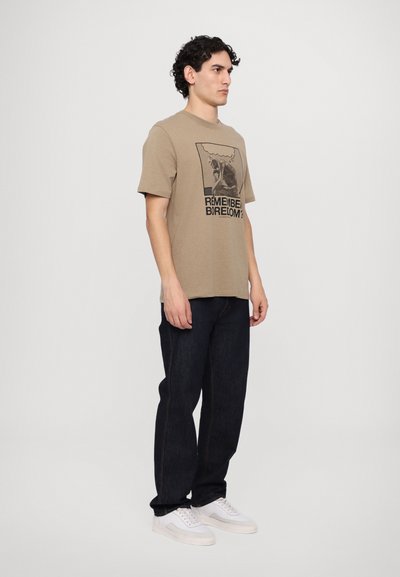 T-shirt en coton beige avec un imprimé graphique portant le texte "TE SOUVIENS-TU DE L'ENNUI ?", associé à un jean en denim bleu foncé et des baskets blanches.