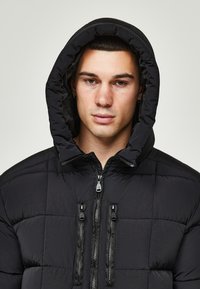 Schwarze gepolsterte Jacke mit Kapuze, mit einem gesteppten Muster, zwei vorderen Reißverschlüssen und zwei RV-Taschen.