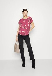 Ružičasta cvjetna bluza s kratkim rukavima, spojena s crnim trapericama i gležnjačama. Model drži bež torbu. Jednostavan, moderan dizajn.