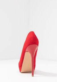 Chaussure à talon haut rouge avec un bout pointu. Texture en daim doux, talon aiguille fin et semelle beige, mettant en valeur un design épuré.