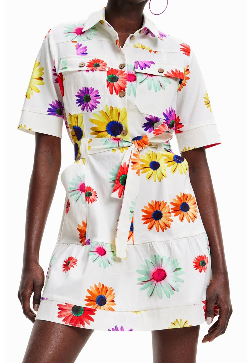 Robe blanche ceinturée avec de grandes impressions florales colorées, manches courtes, poches poitrine boutonnées, portée par une personne à la peau foncée.