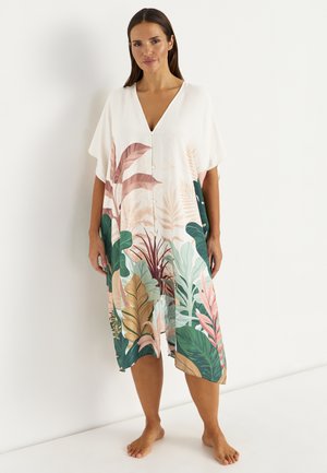 Kobieta stoi boso, ubrana w białą sukienkę do kolan z kolorowym tropikalnym wzorem liści i guzikami z przodu.