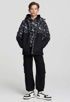 Ragazzo adolescente in piedi con le mani nelle tasche, indossa una giacca marmorizzata bianca e nera, pantaloni cargo neri e sneakers bianco e nere su uno sfondo grigio.