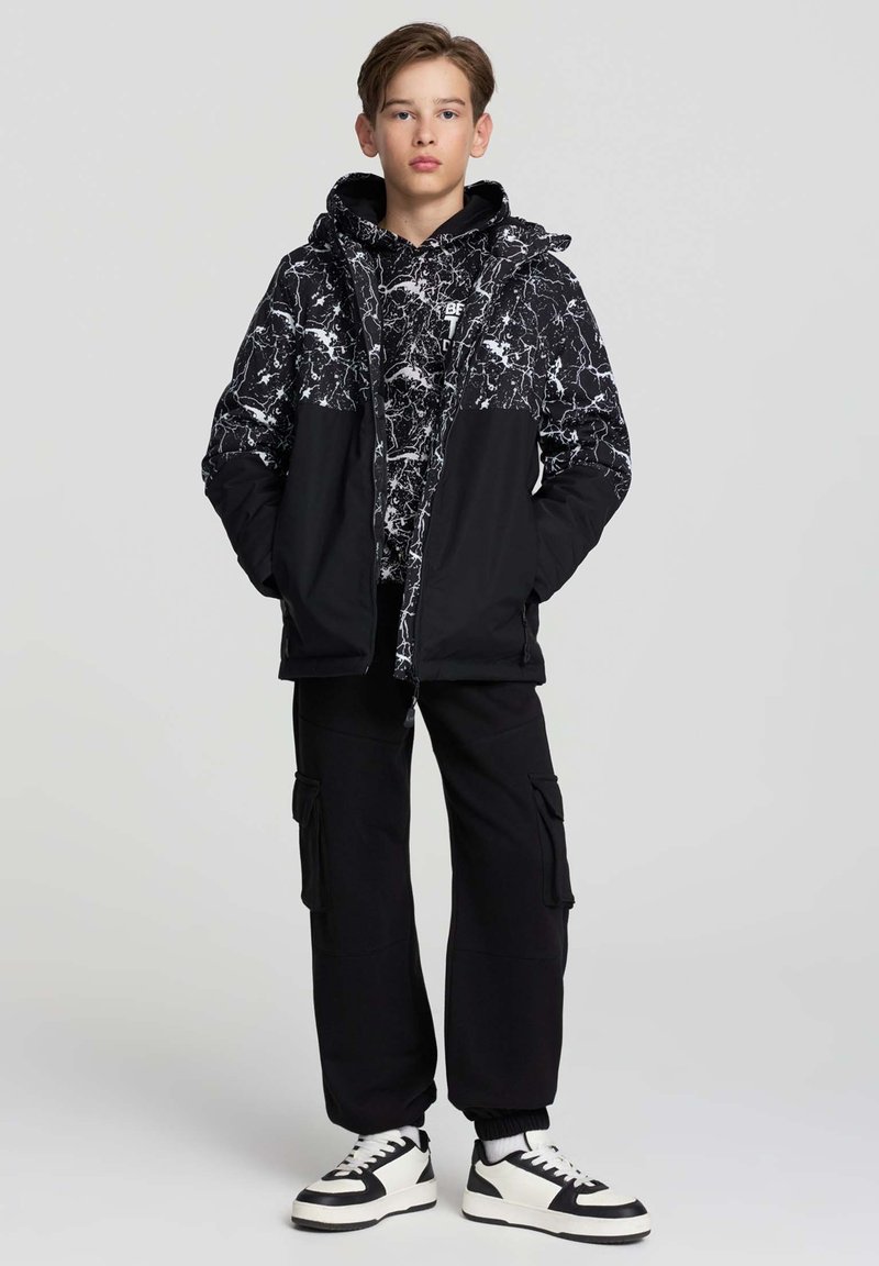 Nastolatek stojący z rękami w kieszeniach, ubrany w czarno-białą marmurkową kurtkę, czarne spodnie cargo, oraz czarno-białe trampki na szarym tle.