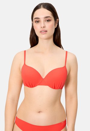 Mujer con cabello largo y castaño que lleva un sujetador rojo ajustado y braguitas de bikini a juego, sobre un fondo liso y claro.