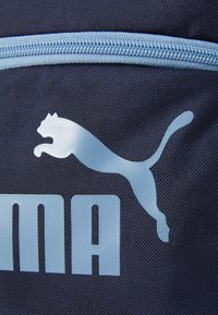Marinblått tyg med en ljusblå Puma-logotyp som visar en hoppande katt och en del av texten "Puma" nedanför logotypen. Dragkedjedetalj synlig högst upp.