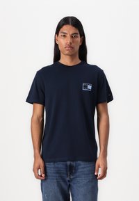 T-shirt en coton bleu marine avec un col rond, des manches courtes et un logo rectangulaire sur la poitrine. Tissu texturé. Design décontracté.