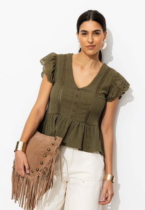 Femme portant un chemisier vert olive texturé avec des manches à œillets, pantalon blanc, bracelets manchettes dorés, tenant un sac à main en suède marron à franges.