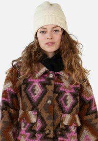 Brown patterned wool coat with pink and cream geometric designs, featuring large buttons and two front pockets. Cream ribbed beanie hat.