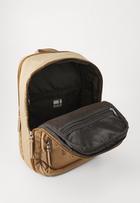 Beige Rucksack aus glattem Stoff, mit einem großen offenen Fach und einer kleineren braunen Reißverschlusstasche, die ein einfaches Design zeigt.