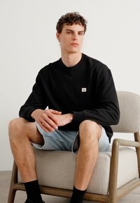 Zwarte sweatshirt met ronde hals en klein logo op de borst, gecombineerd met lichtblauwe denim shorts. Model zittend op een lichtgekleurde stoel.