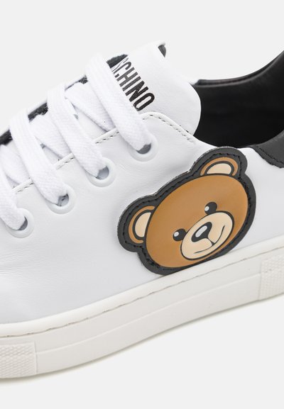 Bílé kožené tenisky s černými akcenty, s gumovou podrážkou a grafikou hnědého medvěda na boku, a bílými tkaničkami.