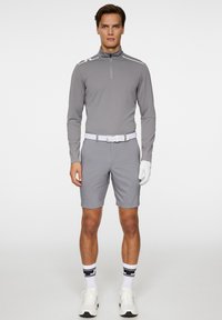 Harmaa pitkähihainen polo, jossa on puolivetoketjukaulus, yhdistettynä harmaisiin shortseihin. Varustettu valkoisella vyöllä ja golf-kämmenmyssyllä, jalassa mustavalkoraidalliset sukat.