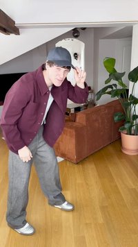 Μπουφάν σε μπορντό χρώμα με κουμπιά, γκρι μπλουζάκι, γκρι παντελόνι και ασημένια παπούτσια. Προσφέρει άνετη εφαρμογή, φορεμένο εσωτερικά με φυτό εσωτερικού χώρου και καναπέ στο φόντο.