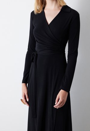 Czarna sukienka kopertowa z długimi rękawami, kołnierzykiem, wiązaniem w talii oraz gładką teksturą. Posiada dopasowany gorset i rozpływającą się spódnicę.